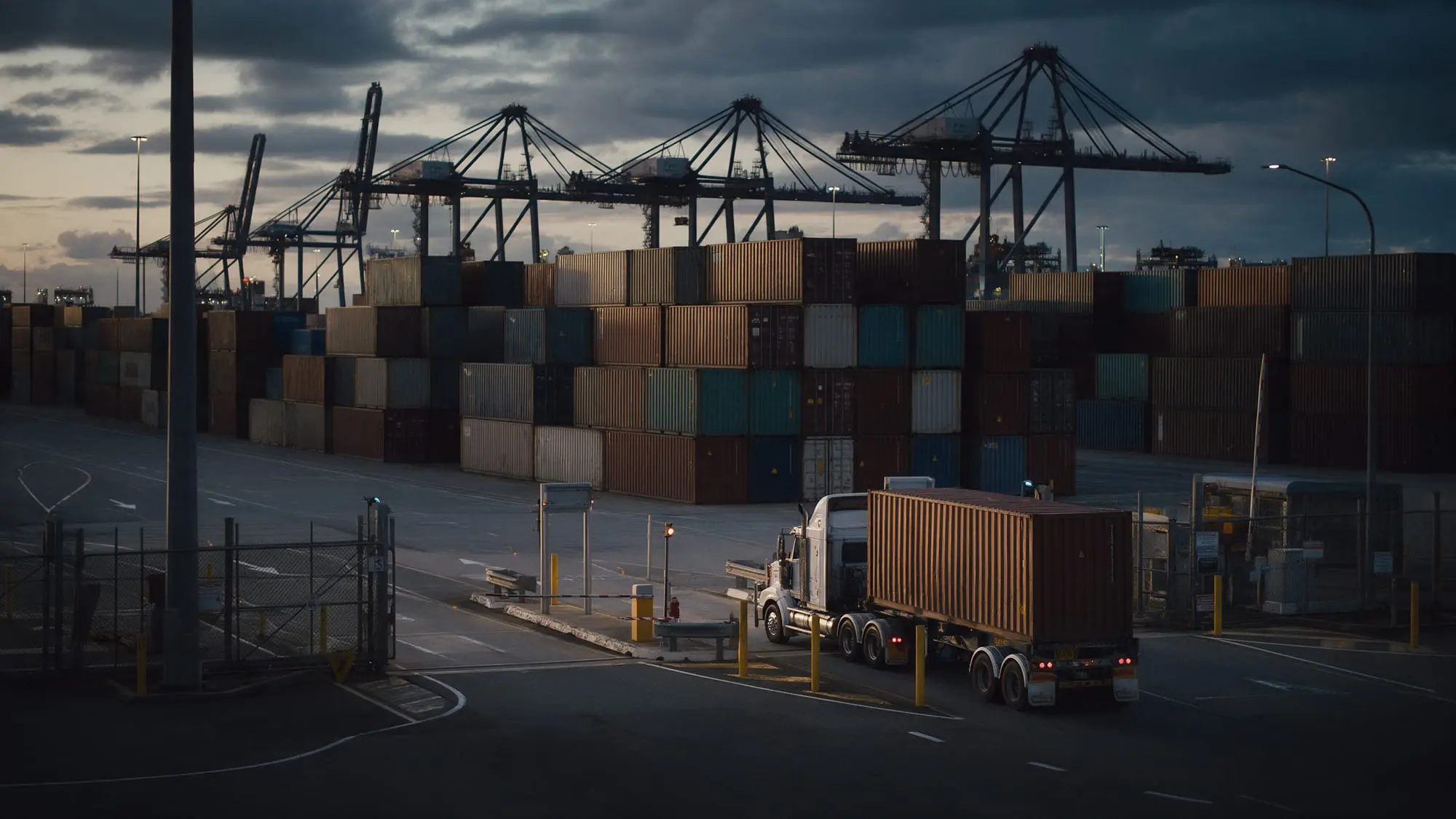 A truck carrying a shipping container past stacked containers and cranes at a port